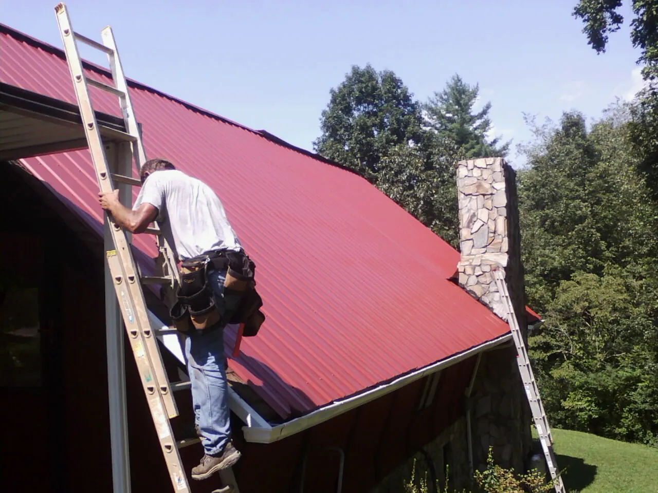 Roofer on a ladder beside a red metal roof and stone chimney — Soffit & Fascia Repair by Roofing Brothers in Cerritos
